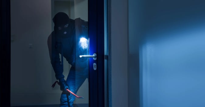 A thief with a flashlight and crowbar trying to open a door.